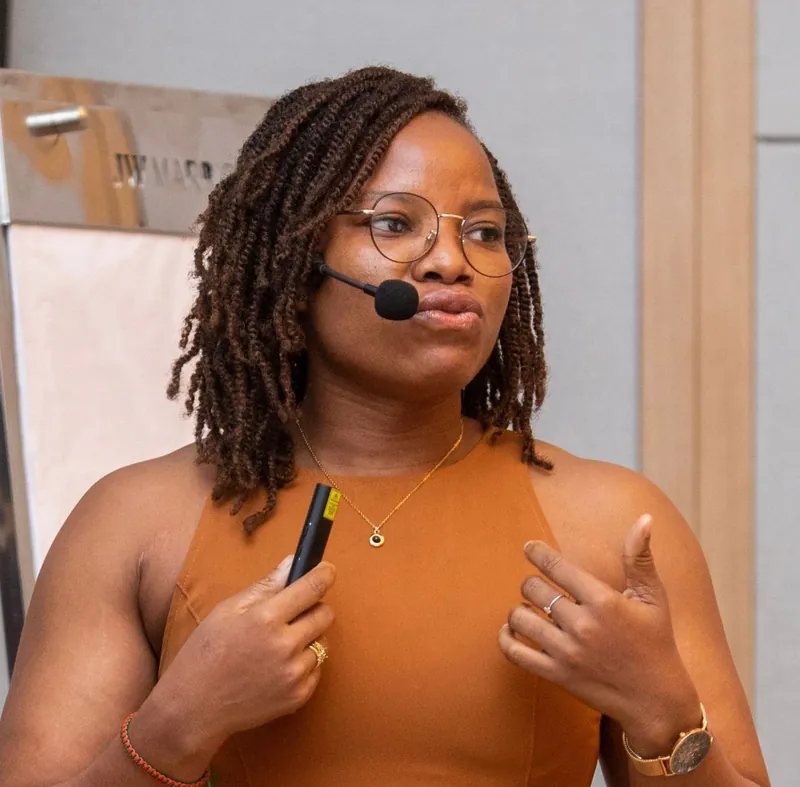 Portrait de Marie Reine Cakpo, en robe orange sans manches et lunettes, micro-casque, en pleine intervention.