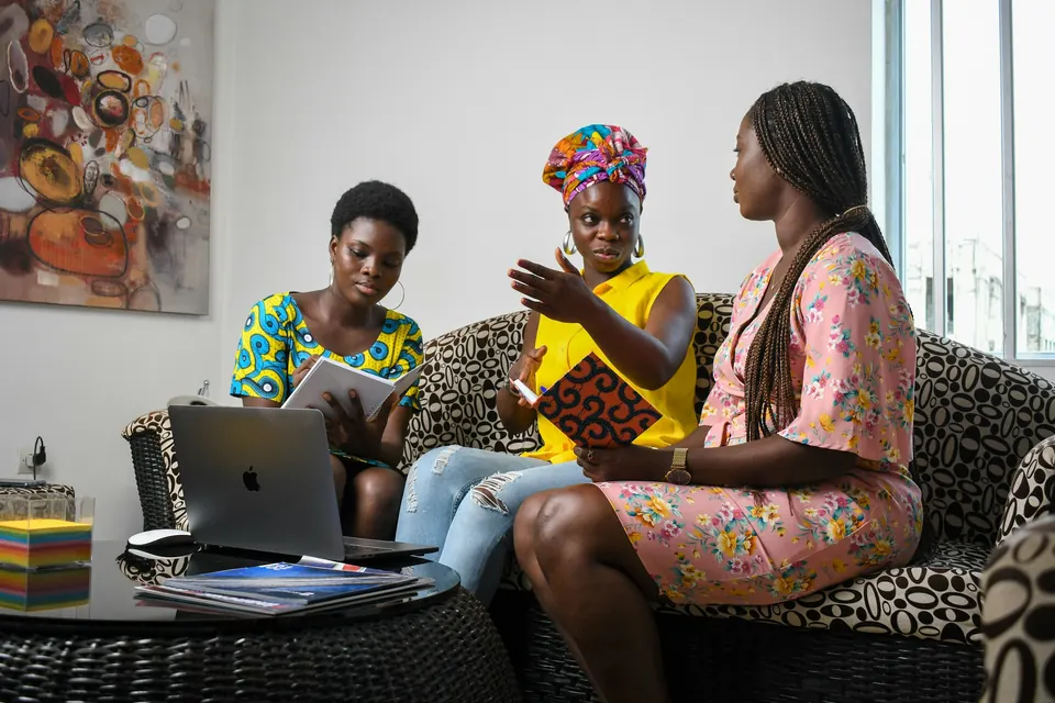 Trois femmes en pleine discussion sur un canapé de bureau, tablette et ordinateur portable à portée de main.